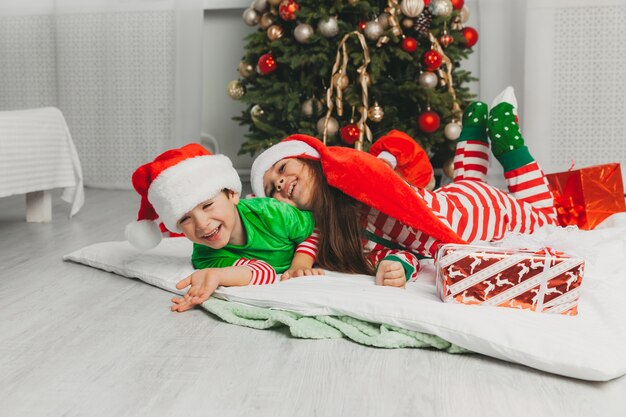 Feliz hermano y hermana vestidos como Santa Claus están sentados cerca del árbol de Navidad en casa en la sala de estar con regalos. Navidad
