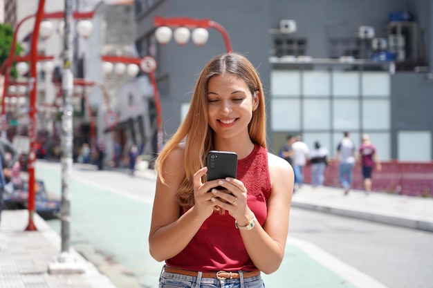 Feliz hermosa adolescente sonriente charlando con su celular caminando en Sao Paulo Brasil