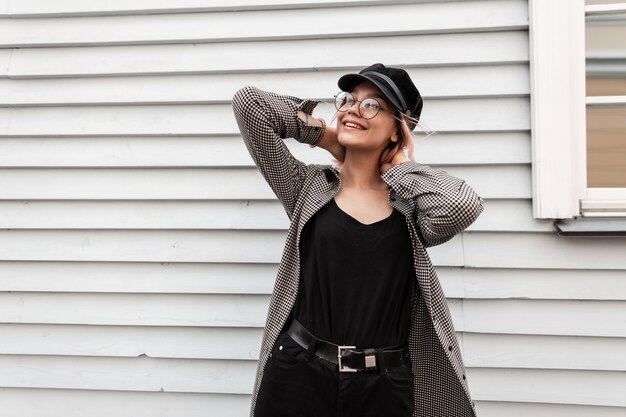 Foto feliz hermosa adolescente sonriente con gafas vintage en ropa de otoño casual de moda se encuentra cerca de una casa de madera