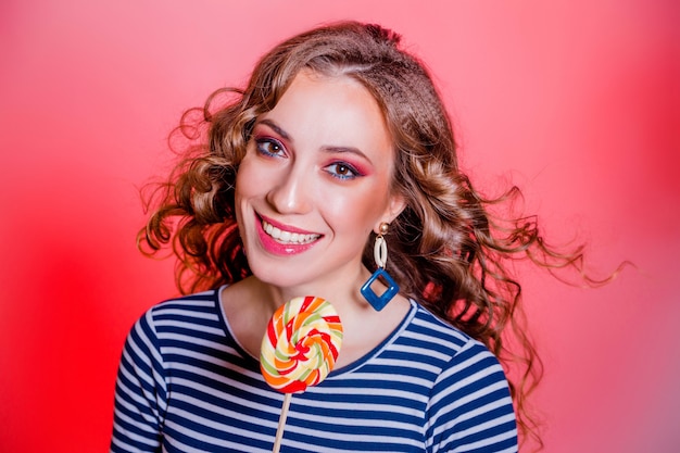 Feliz hermosa chica morena con maquillaje rojo, pelo rizado y un suéter de rayas azules posando sobre un fondo rojo. Foto horizontal