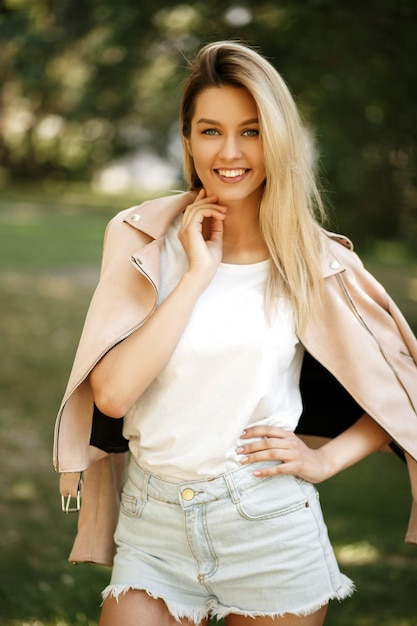 Feliz hermosa chica rubia con una sonrisa en una chaqueta rosa con pantalones cortos en la naturaleza