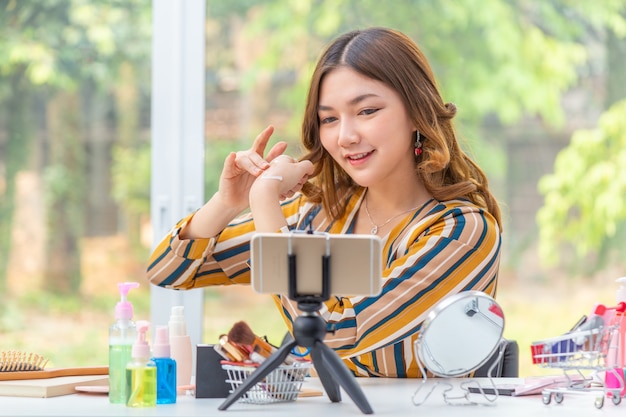 Feliz hermosa joven asiática, vlogger, revisando productos de belleza a través de su video blog en línea desde su casa