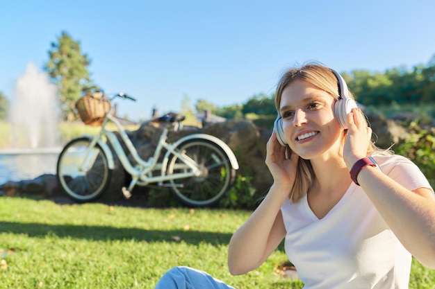 Feliz hermosa joven en auriculares. Chica adolescente escuchando música, estudiando en línea sentado en la naturaleza en el parque en un día soleado de verano. Estilo de vida educativo de los adolescentes, espacio de copia