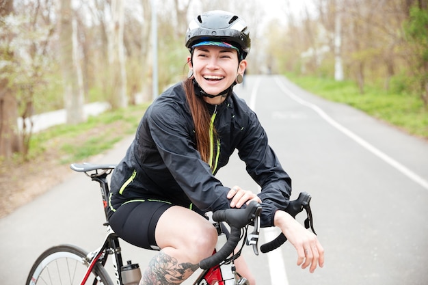 Feliz hermosa joven en casco de bicicleta en bicicleta de pie en la carretera y riendo