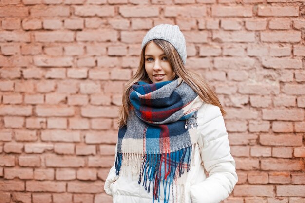 Foto feliz hermosa joven con una chaqueta blanca y un pañuelo de colores cerca de una pared de ladrillos