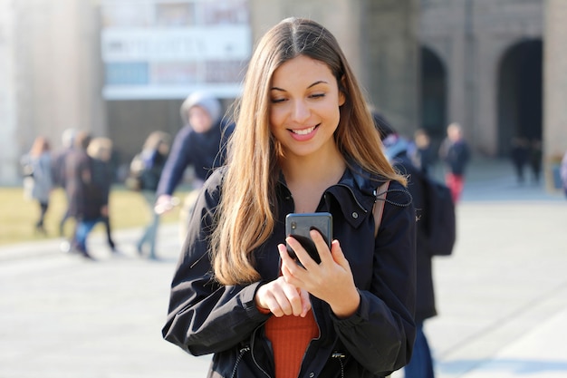 Feliz hermosa joven colegiala con teléfono inteligente al aire libre en día soleado de primavera mensajes de texto y sonriendo