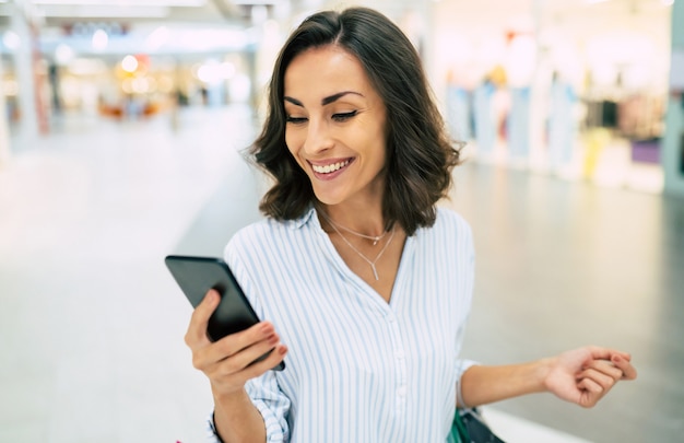 Feliz hermosa joven elegante con bolsas de la compra está usando un teléfono inteligente mientras camina en el centro comercial