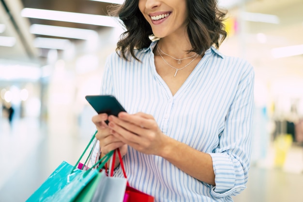 Foto feliz hermosa joven elegante con bolsas de la compra está usando un teléfono inteligente mientras camina en el centro comercial