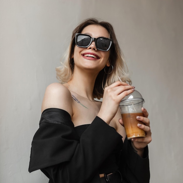 Feliz hermosa joven elegante con gafas de sol de moda en ropa de negocios negra de moda con top y blazer bebiendo café frío con jugo y disfrutando de un paseo al aire libre cerca de la pared blanca