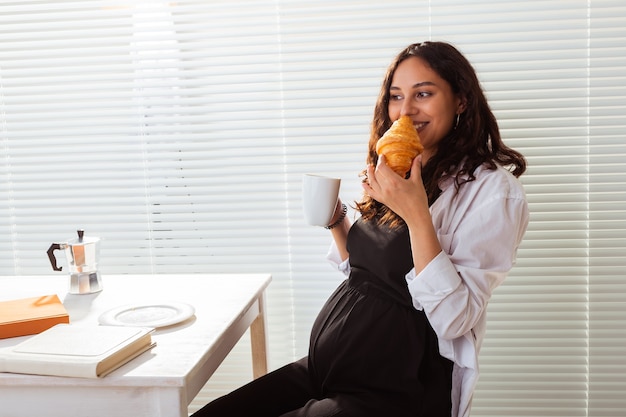 Feliz hermosa joven embarazada comiendo croissant durante el desayuno de la mañana concepto de agradable