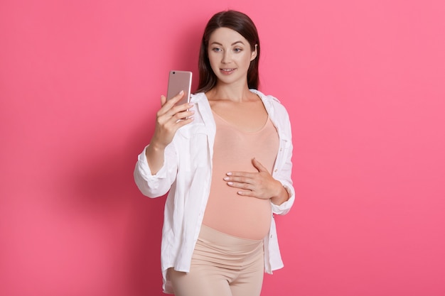 Feliz hermosa joven embarazada toma selfie en su teléfono inteligente sobre fondo rosa, vistiendo, camisa y leggins, manteniendo la palma en su vientre, mira la pantalla del dispositivo.