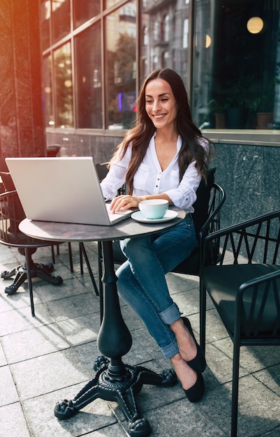 Feliz hermosa joven empresaria trabajando en una laptop en un café callejero al aire libre