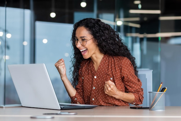 Feliz hermosa joven latinoamericana sentada en la oficina frente al monitor de la computadora portátil