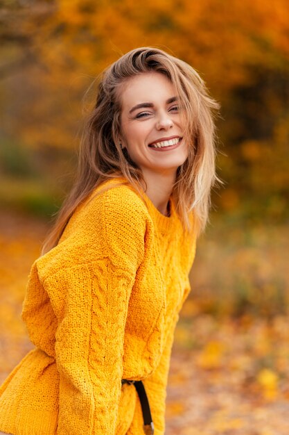 Feliz hermosa joven con una linda sonrisa en un suéter de punto amarillo en un parque de otoño con hojas de otoño dorado brillante. Emociones positivas femeninas al aire libre