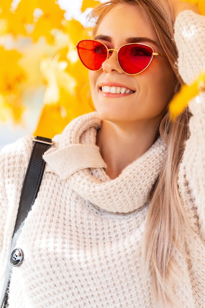 Feliz hermosa joven con una linda sonrisa en un suéter de punto de moda con gafas rojas y una mochila caminando en un parque de otoño con hojas coloridas y brillantes