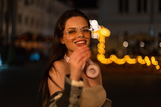 Feliz hermosa joven modelo con una sonrisa en ropa de moda con gafas y auriculares camina en la ciudad nocturna con luces bokeh