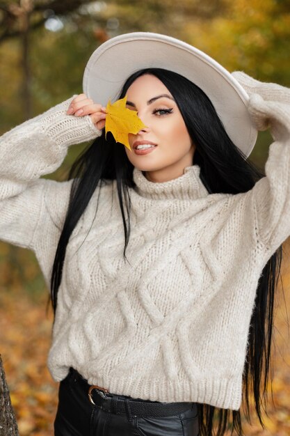 Feliz hermosa joven modelo con sonrisa en suéter de punto blanco y sombrero sostiene una hoja amarilla de otoño y camina en el colorido parque de otoño