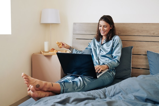 Feliz hermosa joven pasar el fin de semana en casa, comer croissants y ver series en la computadora portátil