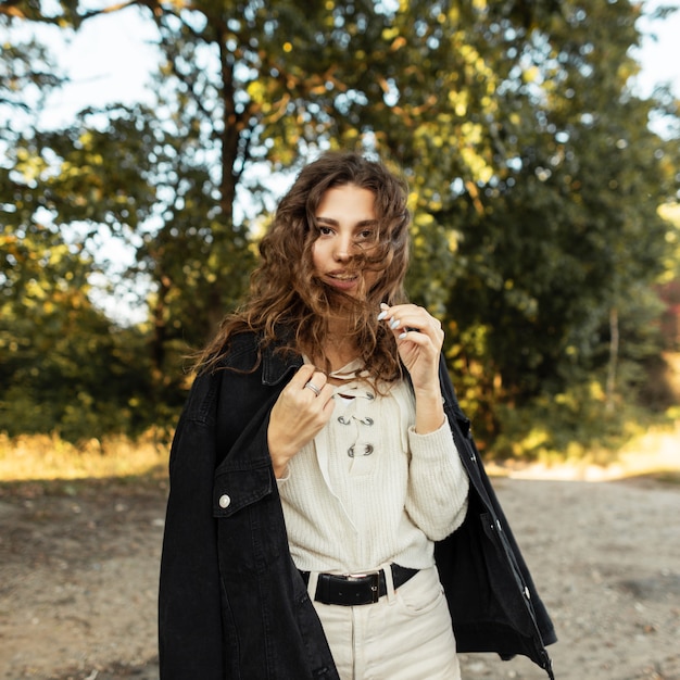 Feliz hermosa joven con el pelo rizado en una chaqueta de mezclilla de moda con una blusa tejida en la naturaleza en el fondo de los árboles