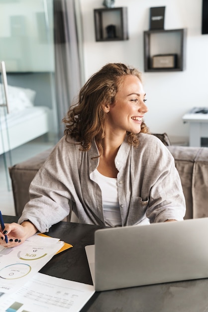 Feliz hermosa joven rubia mujer sentada en la mesa en casa, trabajando en la computadora portátil, tomando notas