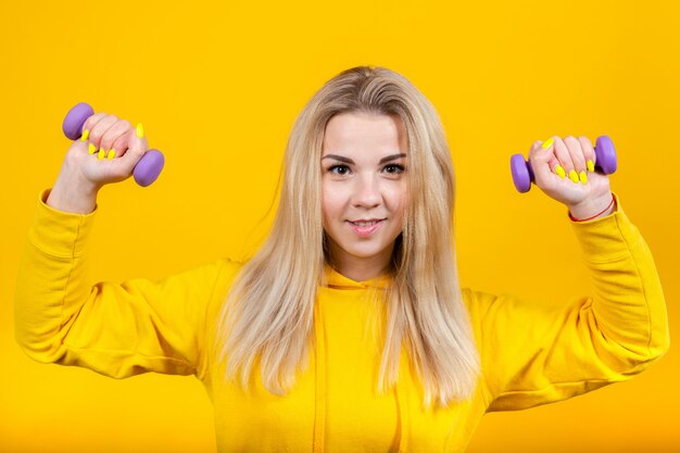 Foto feliz hermosa joven rubia en ropa deportiva amarilla casual haciendo ejercicios con pequeñas pesas moradas de 0.5 kg.