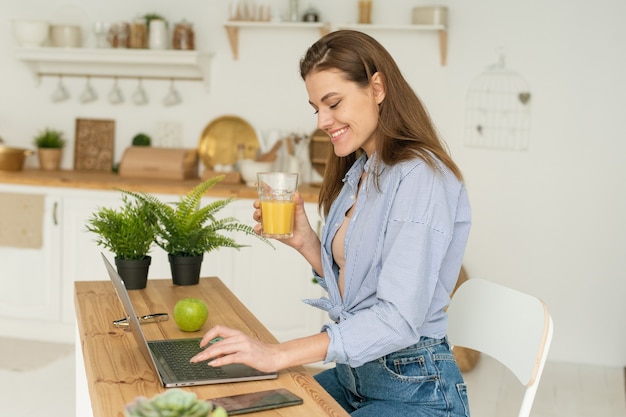 Feliz hermosa joven sentada a la mesa en casa, trabajando en una computadora portátil y bebiendo un delicioso jugo de naranja. Trabajar y estudiar desde casa en condiciones de cuarentena.