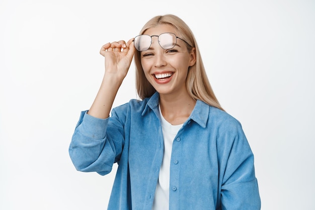 Foto feliz hermosa modelo femenina quitándose las gafas sonriendo y mirando alegre a la cámara de pie con ropa informal sobre fondo blanco.