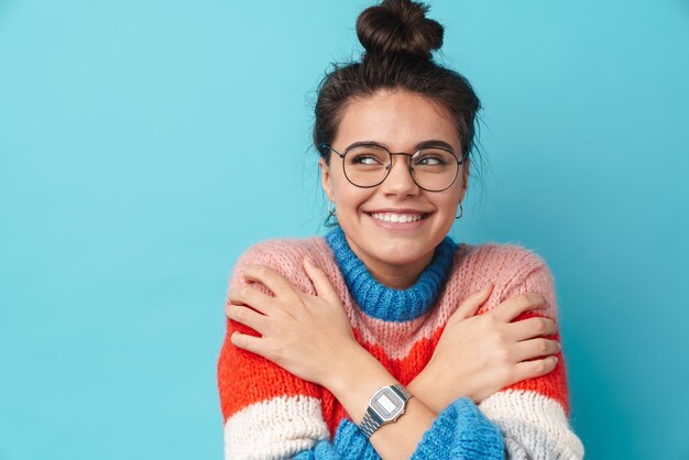 Feliz hermosa mujer en anteojos sonriendo y abrazándose a sí misma aislada sobre pared azul