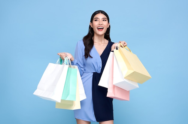 Feliz hermosa mujer asiática adicta a las compras con vestido azul y sosteniendo bolsas de compras aisladas en la pared azul.