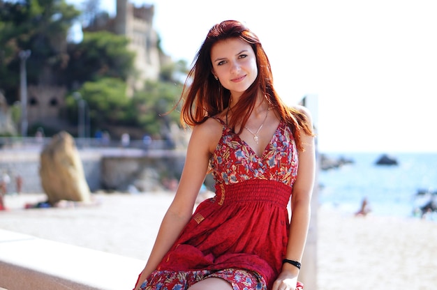 Feliz hermosa mujer de cabello rojo con linda sonrisa en vestido rojo en la playa