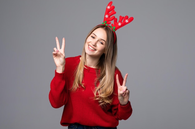 Feliz hermosa mujer con cuernos de reno en la cabeza mira con gesto de paz en la pared gris