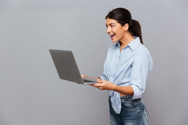 Feliz hermosa mujer emocionada usando laptop aislado en una pared gris