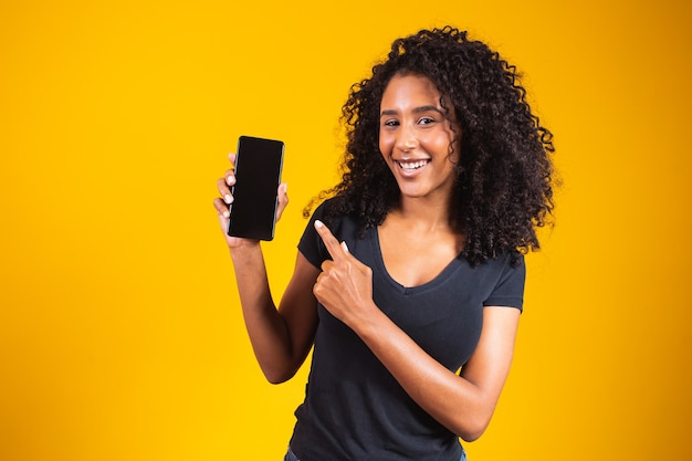 Feliz hermosa mujer joven con cabello afro sosteniendo teléfono celular de pantalla en blanco y dedo acusador sobre fondo amarillo.