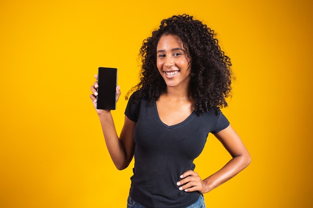 Feliz hermosa mujer joven con cabello afro con teléfono celular de pantalla en blanco sobre fondo amarillo.