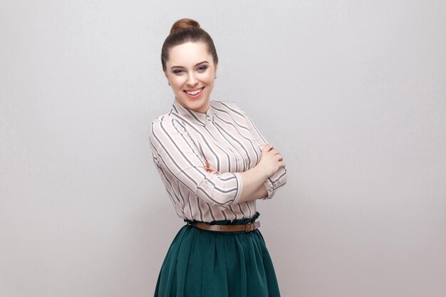 Feliz hermosa mujer joven en camisa a rayas, falda verde con maquillaje y peinado de prohibición, de pie con las manos cruzadas y mirando a la cámara con una gran sonrisa. tiro del estudio, aislado en fondo gris.