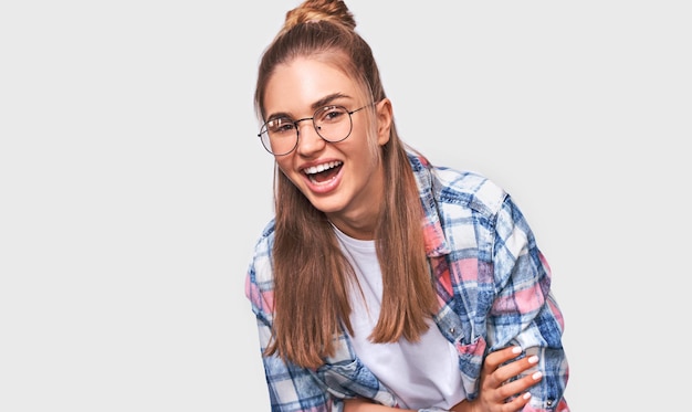 Feliz hermosa mujer joven positiva vestida con ropa informal con anteojos transparentes redondos con una amplia sonrisa agradable mirando a la cámara y posando sobre la pared blanca del estudio Emociones de la gente