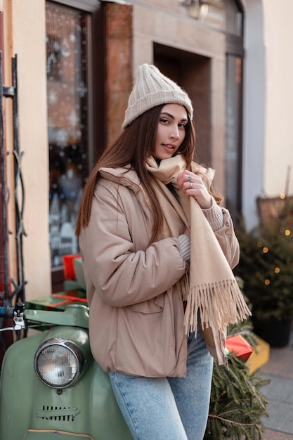 Feliz hermosa mujer joven en ropa de invierno de moda con un sombrero de  punto, chaqueta, bufanda y jeans se encuentra cerca de una moto con árboles  de navidad y regalos en