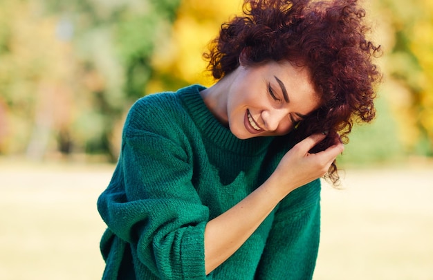 Feliz hermosa mujer joven sonriendo con la mano en el cabello contra el fondo de la naturaleza con el pelo rizado ventoso tiene una expresión positiva vestida con un suéter verde La gente viaja y el concepto de estilo de vida