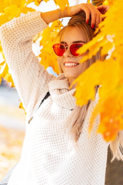 Feliz hermosa mujer joven con una sonrisa y gafas de sol rojas con un suéter de punto blanco de moda camina en un parque de otoño con hojas amarillas y disfruta de un cálido día de otoño