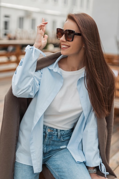 Foto feliz hermosa mujer joven con una sonrisa con gafas de sol en ropa casual de mezclilla de moda con una camisa de abrigo y jeans se divierte en la ciudad de primavera belleza de estilo y apariencia de moda