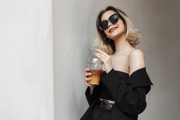 Feliz hermosa mujer joven con una sonrisa y gafas de sol con ropa elegante negra de moda caminando por la calle cerca de la pared blanca y bebiendo café frío Chica bebe y disfruta