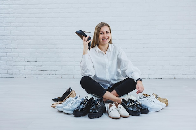 Feliz hermosa mujer joven con una sonrisa en un traje de moda con una camisa blanca y pantalón negro se sienta en el suelo entre los zapatos y elige un nuevo par de zapatos para ella