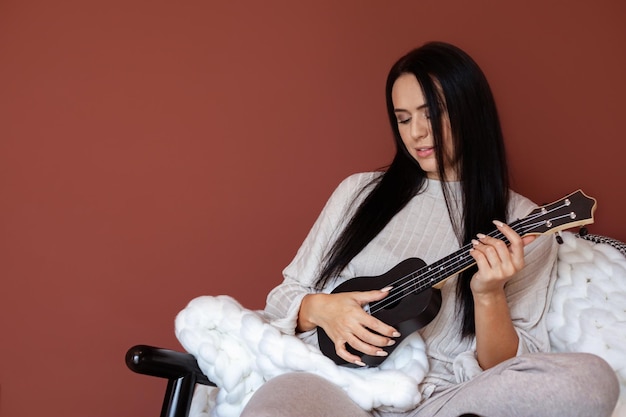 Feliz hermosa mujer músico tocando el ukelele escribiendo música sentado en un cómodo sofá en casa