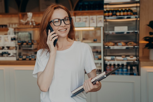 Feliz hermosa mujer de negocios hablando por teléfono celular fuera de pie con el portátil cerca de la cafetería