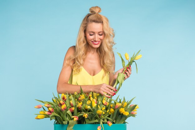 Feliz hermosa mujer rubia caucásica con caja grande de tulipanes sobre fondo azul.