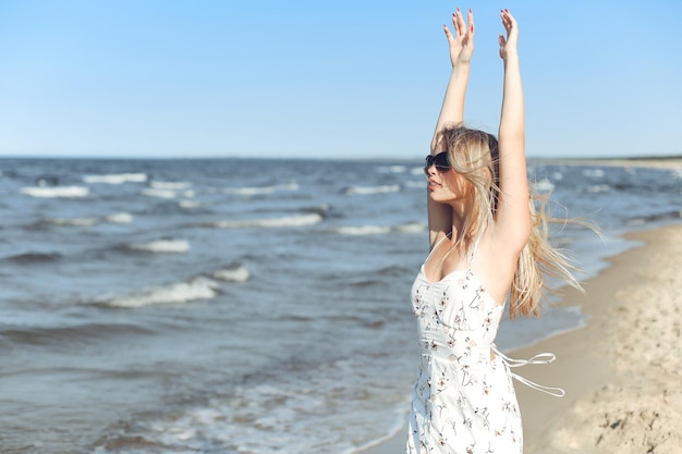 Feliz hermosa mujer rubia en la playa del océano de pie con un vestido blanco de verano y gafas, levantando las manos.