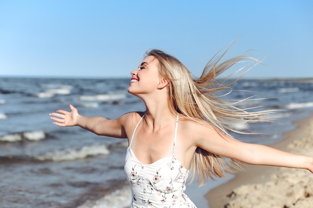 Feliz hermosa mujer rubia en la playa del océano de pie con un vestido blanco de verano, levantando las manos.