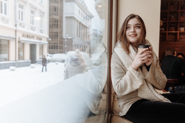 Feliz hermosa mujer en suéter blanco y bufanda sentada junto a la ventana en el café