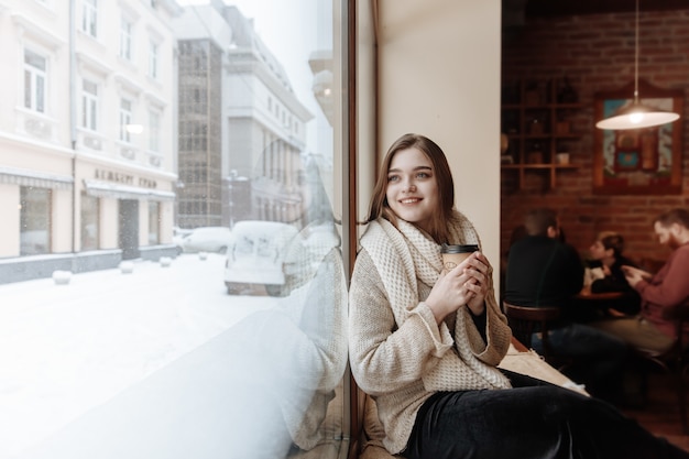 Feliz hermosa mujer en suéter blanco y bufanda sentada junto a la ventana en el café