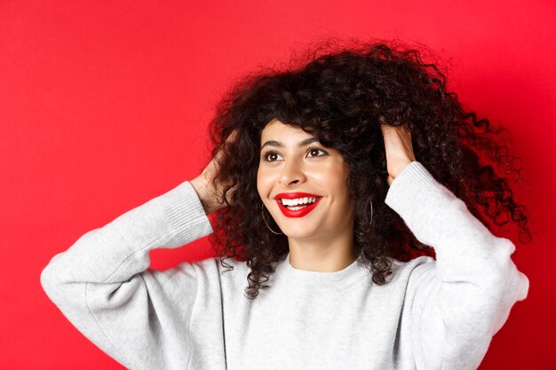 Feliz hermosa mujer tocando el cabello rizado natural y sonriendo complacido, mirando a la izquierda, como su peinado, de pie contra el fondo rojo.